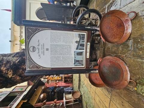       Informative signboard for Medersa Seffarine displayed on a rainy street with copper trays at the base.
  