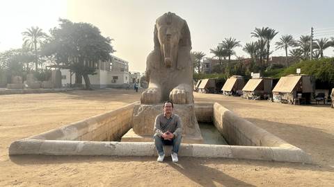       A traveller sits cross-legged before the alabaster Sphinx of Memphis on a hazy morning.
  