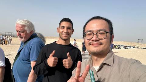       Travellers smile and gesture for a selfie in the desert with distant tour groups behind.
  