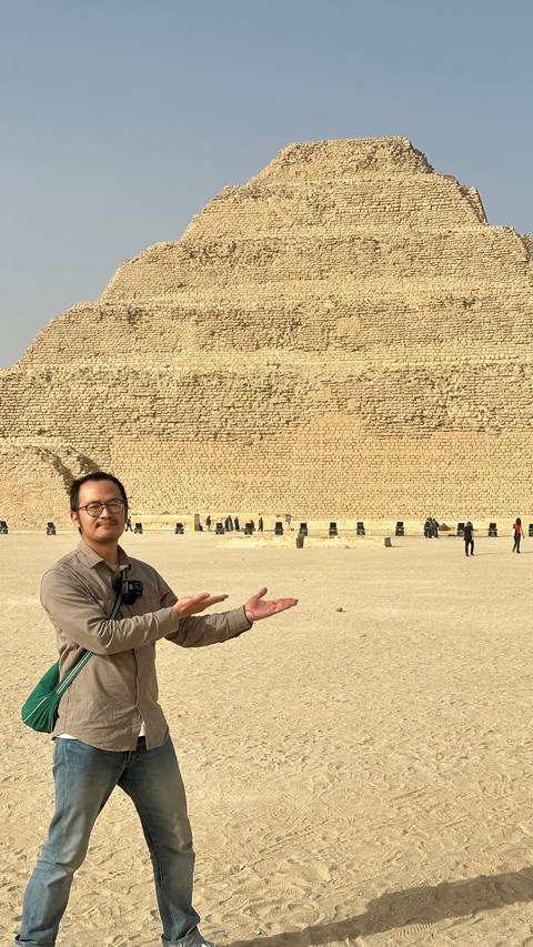      A smiling tourist stands in the desert framing the Great Pyramid of Giza with his hands.
  