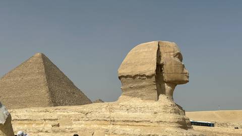       Side profile of the Great Sphinx with the Pyramid of Khafre rising behind it under a blue sky.
  