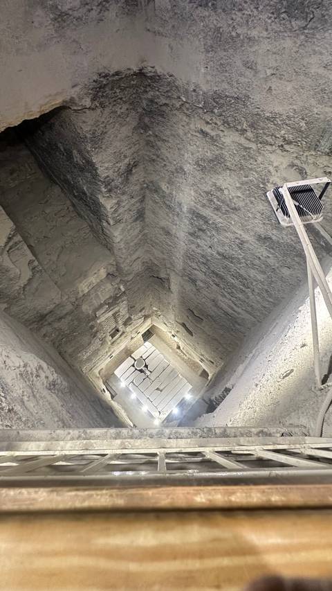       Dimly lit sloping interior passageway inside an ancient pyramid with rough stone walls and a ventilator fan.
  