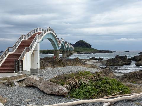       Arched Sanxiantai footbridge spans rocky shoreline toward a small offshore island under gray skies.
  