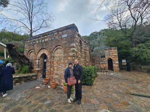       Couple poses before the stone house said to be the last home of the Virgin Mary, surrounded by greenery.
  