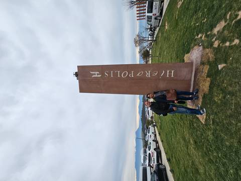       Visitors stand beside a tall brown stone monument marked 'Hierapolis' on a grassy plain.
  
