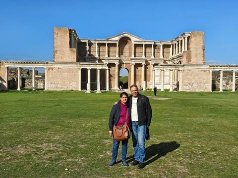      Couple stands on grassy forecourt before the grand colonnaded façade of the ancient Sardis gymnasium.
  
