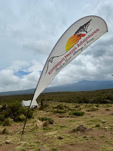       Flag bearing Kilimanjaro Heroes logo fluttering above a campsite with Mount Kilimanjaro in the distance
  