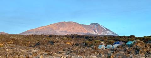       Tents pitched on rocky terrain beneath snow-tipped Mount Kilimanjaro at sunrise
  