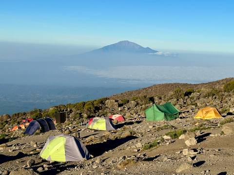       Colorful tents scattered on a rocky alpine plateau with a distant volcanic peak rising above the plains.
  