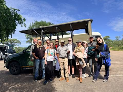       Tour group and guide smile beside an open-sided green safari vehicle under a blue sky.
  