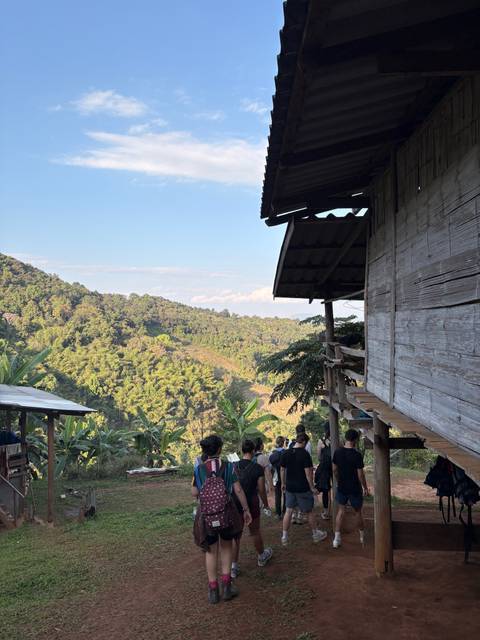       Verdant mountain valley viewed from a rustic wooden hut balcony with travelers relaxing below.
  