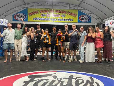       Large tour group and two Muay Thai fighters pose together inside a boxing ring at a night market arena.
  