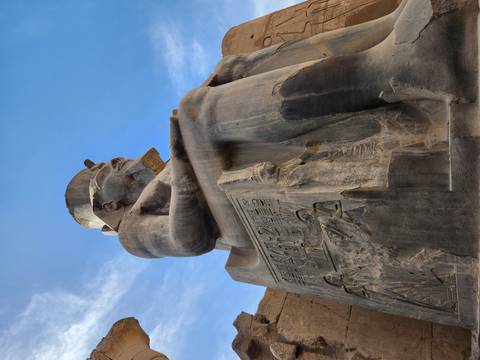       Low-angle view of a colossal granite statue of a seated pharaoh against a bright sky.
  