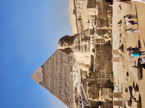       Iconic view of the Sphinx with the Pyramid of Khafre towering behind and visitors below.
  