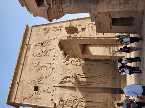      Travelers entering the monumental pylon richly carved with Egyptian deities at Edfu Temple.
  