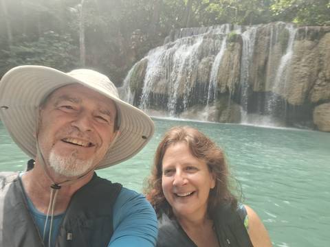       Selfie of an older couple standing before a turquoise pool and tiered waterfall in bright daylight.
  