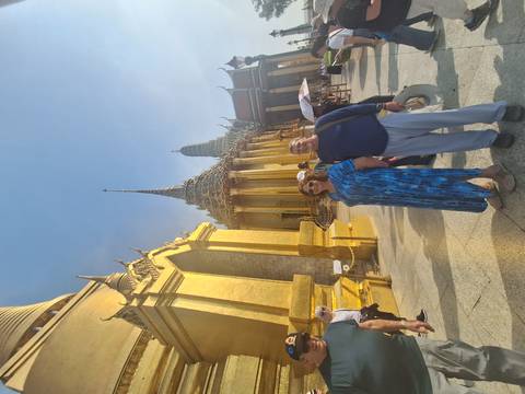       Visitors pose in front of the gleaming golden Grand Palace complex with ornate spires in Bangkok.
  