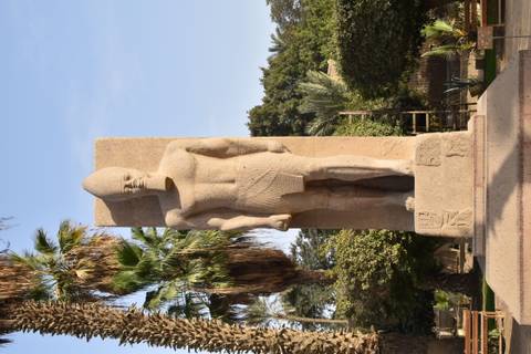       Large statue of Ramses II standing amidst palm trees in an open museum garden.
  