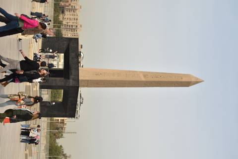       Tall ancient obelisk surrounded by crowds at a modern plaza.
  