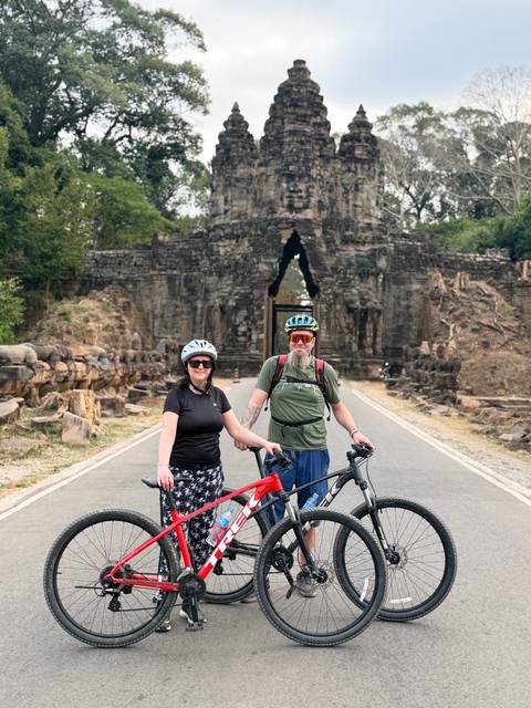       Cyclists pose with bikes on road leading to ancient Angkor stone gate lined with statues.
  