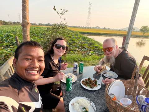       Three cyclists enjoy outdoor meal beside lush rice fields and lotus pond at sunset.
  
