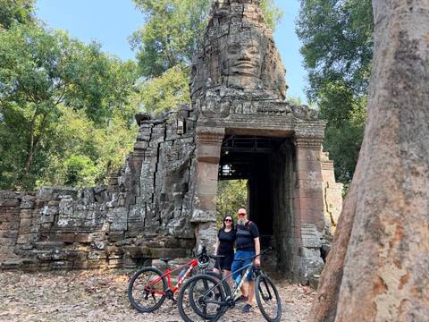       Cyclists stand beside bike at stone face tower entrance in Angkor complex under dappled shade.
  