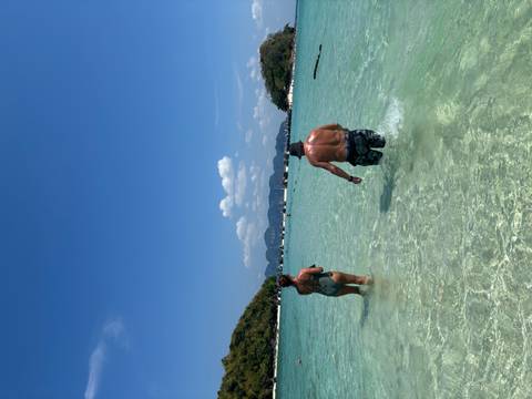       A man and woman wade through clear shallow water toward anchored boats with mountains in the distance.
  