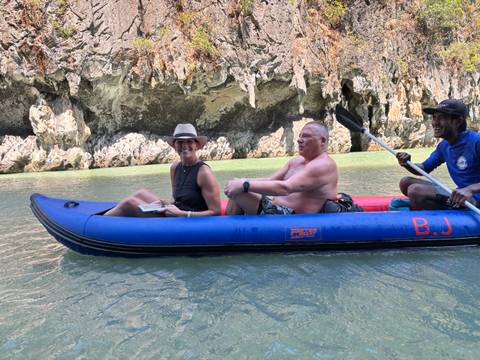       Smiling travellers seated in a blue inflatable canoe gliding along jade-green water beneath limestone cliffs.
  