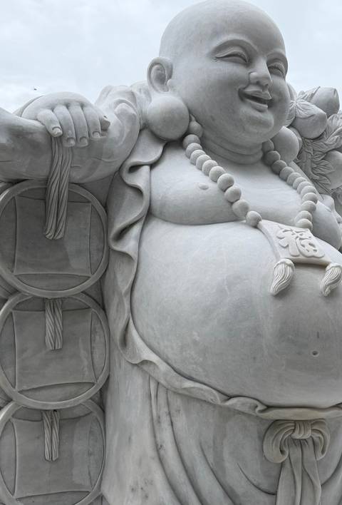       Close-up detail of a large stone laughing Buddha statue with prayer beads.
  