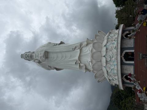       Towering white Lady Buddha statue atop a lotus pedestal against a moody sky.
  