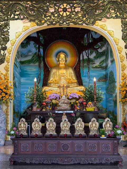       Vibrant altar with golden seated Buddha surrounded by offerings, flowers and murals.
  