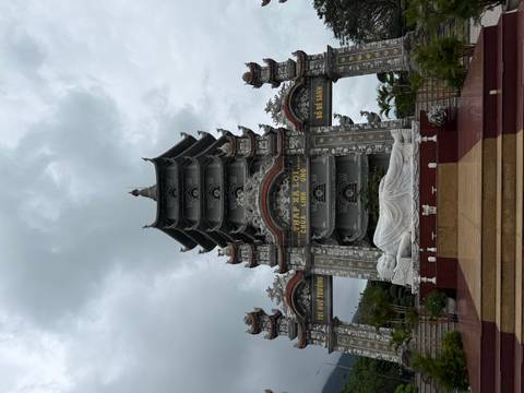       Pagoda complex with reclining white Buddha statue in front of ornate gray tower under cloudy skies.
  