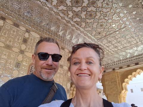       Smiling couple takes a close-up selfie beneath an ornate mirrored mosaic ceiling in an Indian palace.
  