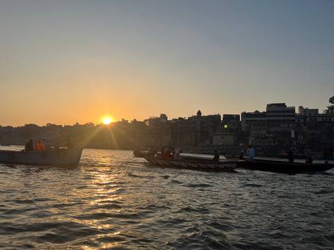       Rowboats carrying passengers move along a shimmering river under a glowing sunset with city silhouettes on the bank.
  