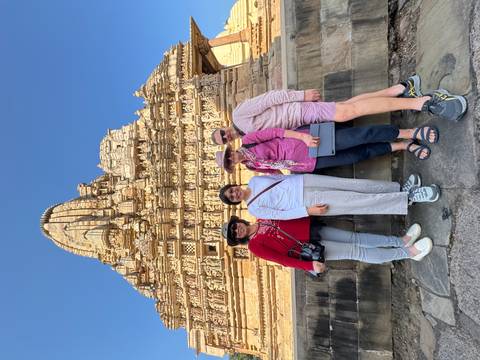       Four travellers pose smiling in front of an intricately carved sandstone temple under a bright blue sky.
  