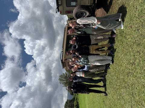       Large group of safari travellers pose in front of an open-roof jeep on grassy plains under partly cloudy skies.
  