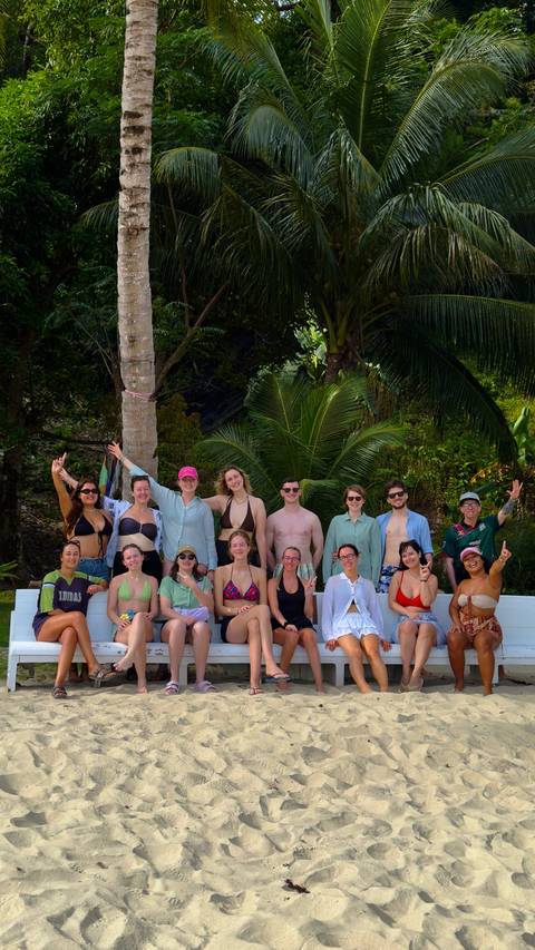       A large group of swimsuit-clad travellers posing together on a bench framed by tropical palm trees and jungle greenery.
  