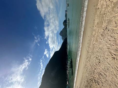       A tranquil tropical beach with soft sand, emerald water, fishing boats and dramatic limestone mountains under a clear blue sky.
  