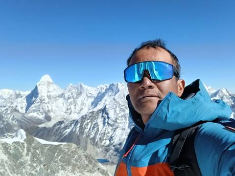       Selfie of a climber in reflective sunglasses with dramatic Himalayan peaks and deep blue sky behind.
  