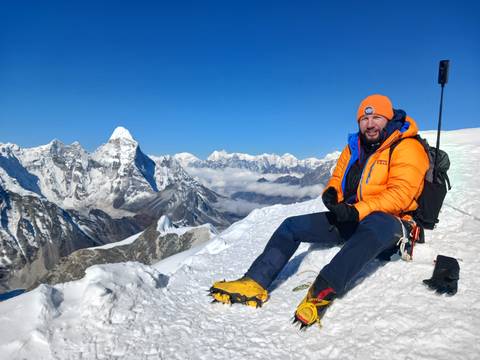       Mountaineer in bright orange jacket and crampons rests on a snowy summit ridge with vast Himalayan panorama.
  