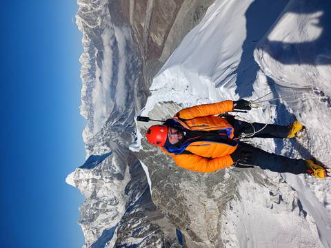       Climber roped in on a snowy ridge wearing helmet and bright jacket with dramatic peaks behind.
  