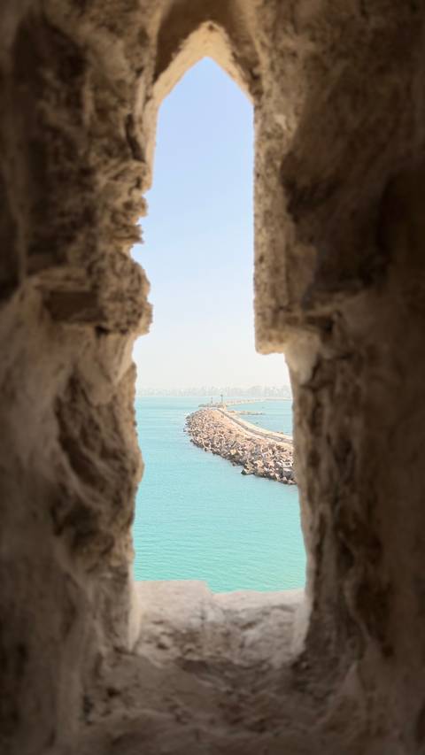      View of a long stone breakwater and distant city skyline framed by weathered castle walls over turquoise water.
  