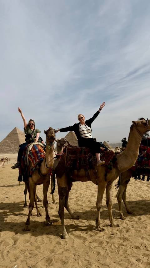       Two travellers joyfully ride camels in front of the iconic Pyramids of Giza under a hazy sky.
  