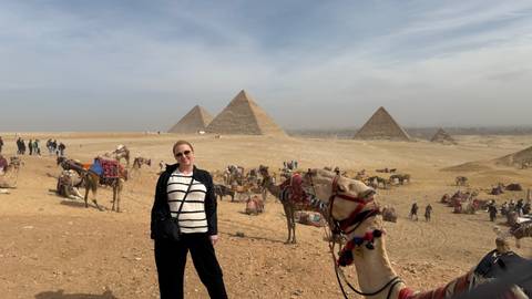       Visitor stands smiling with expansive desert plateau and three Pyramids of Giza rising behind.
  