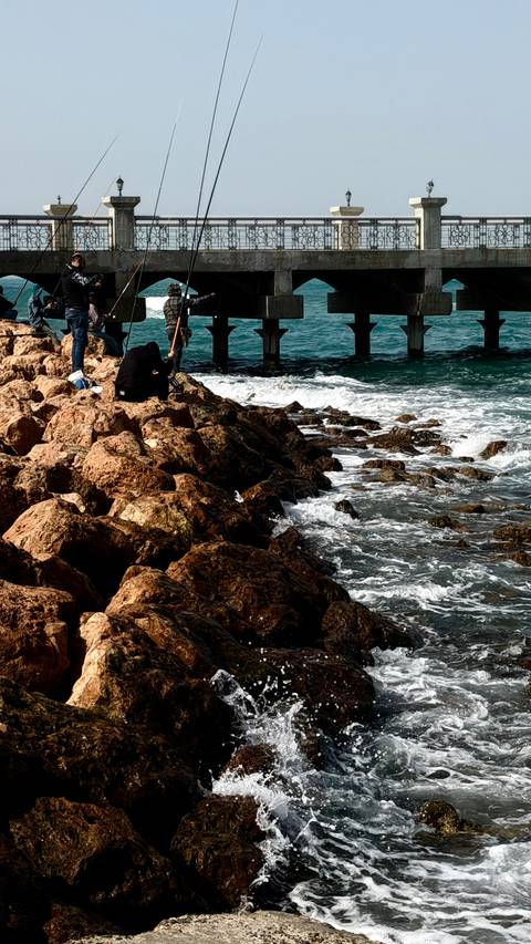      People sit and fish on rocky shore beside turquoise Red Sea with a pier supported by concrete pillars.
  