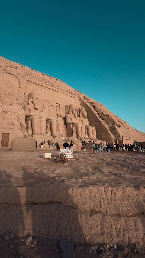      Morning light illuminates the colossal seated statues of Abu Simbel as crowds gather below the temple façade.
  
