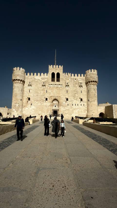       Visitors walk toward a massive stone fortress with twin towers under deep blue sky
  