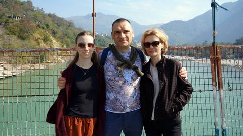       Three travelers pose on a suspension bridge over a turquoise river with mountains beyond
  
