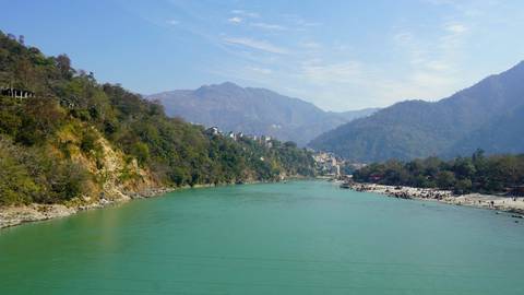       Aqua-blue Ganges flowing through a steep wooded valley beneath a bright sky
  