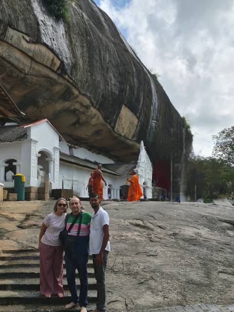       Travellers and robed monks at the entrance of a cave temple built beneath an overhanging rock.
  